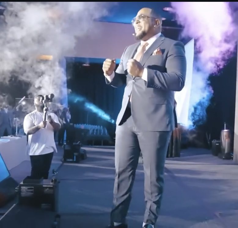 Richard Hall Jr. on stage in a grey suit, fists raised, smoke rising behind him
